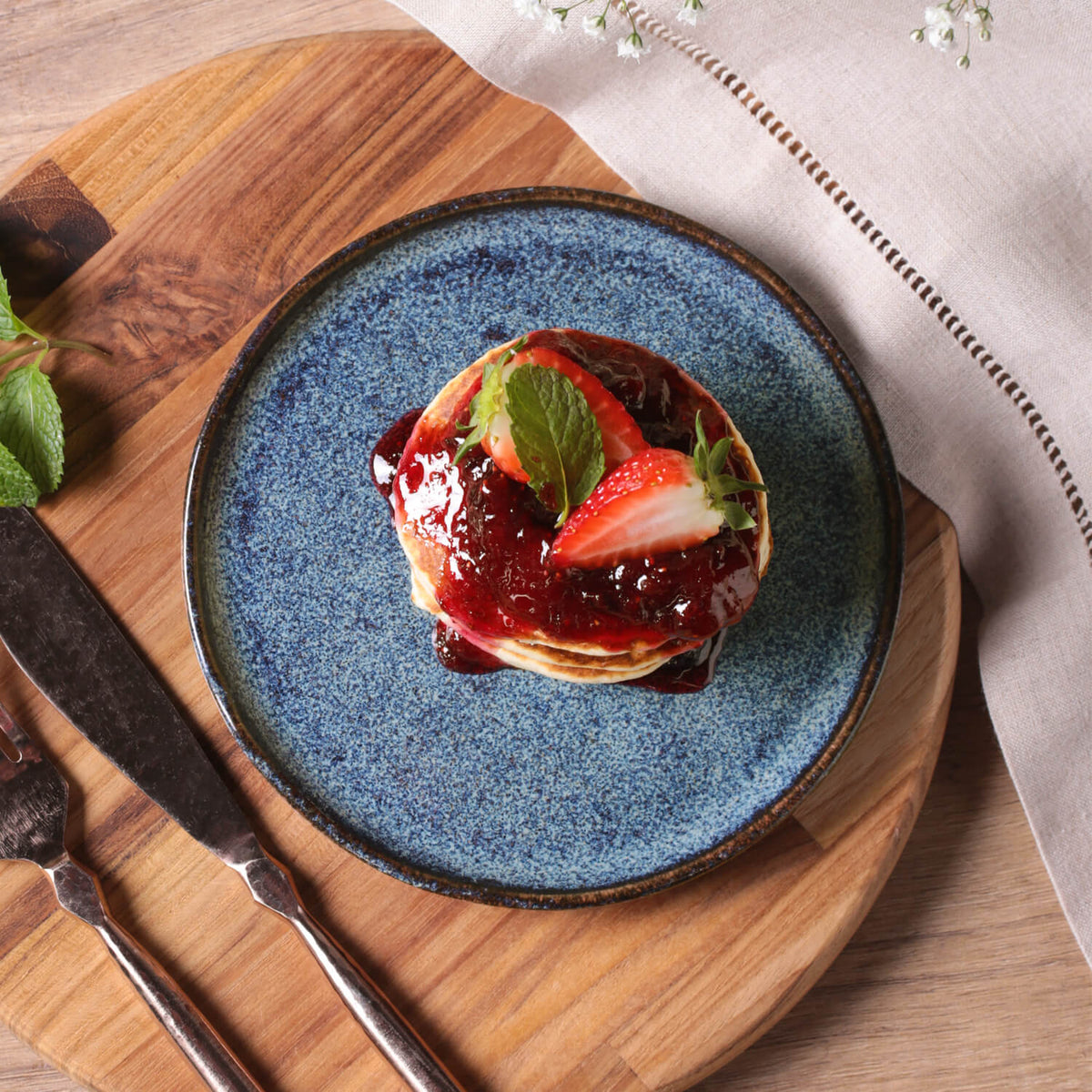 Blue ceramic plate with pancakes topped with strawberry jam and a strawberry on a wooden board.
