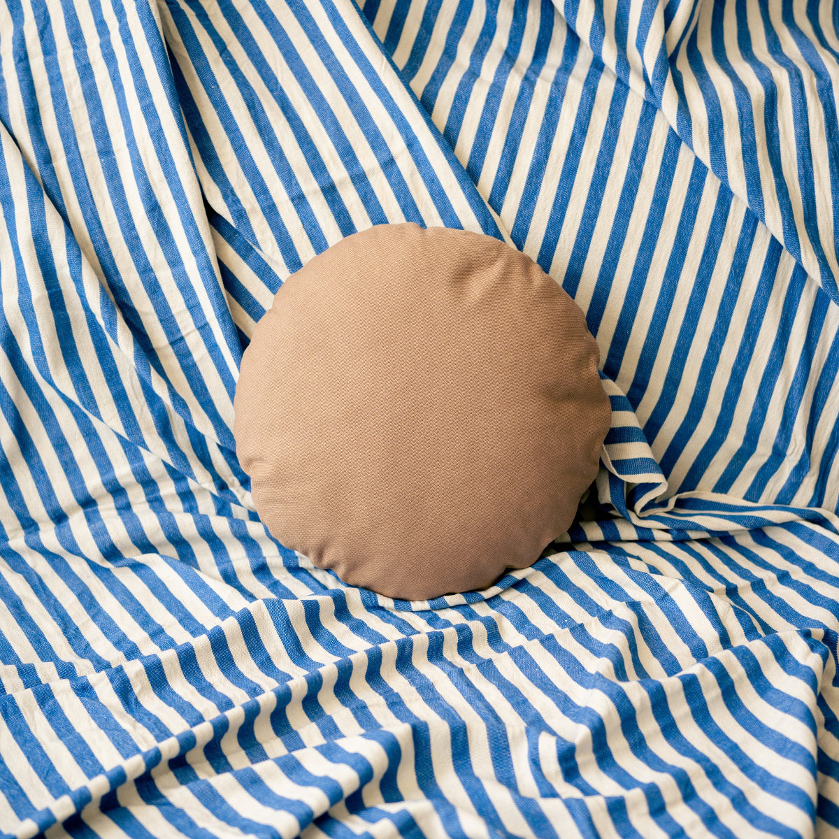 Round light brown pillow on a blue and white striped fabric background