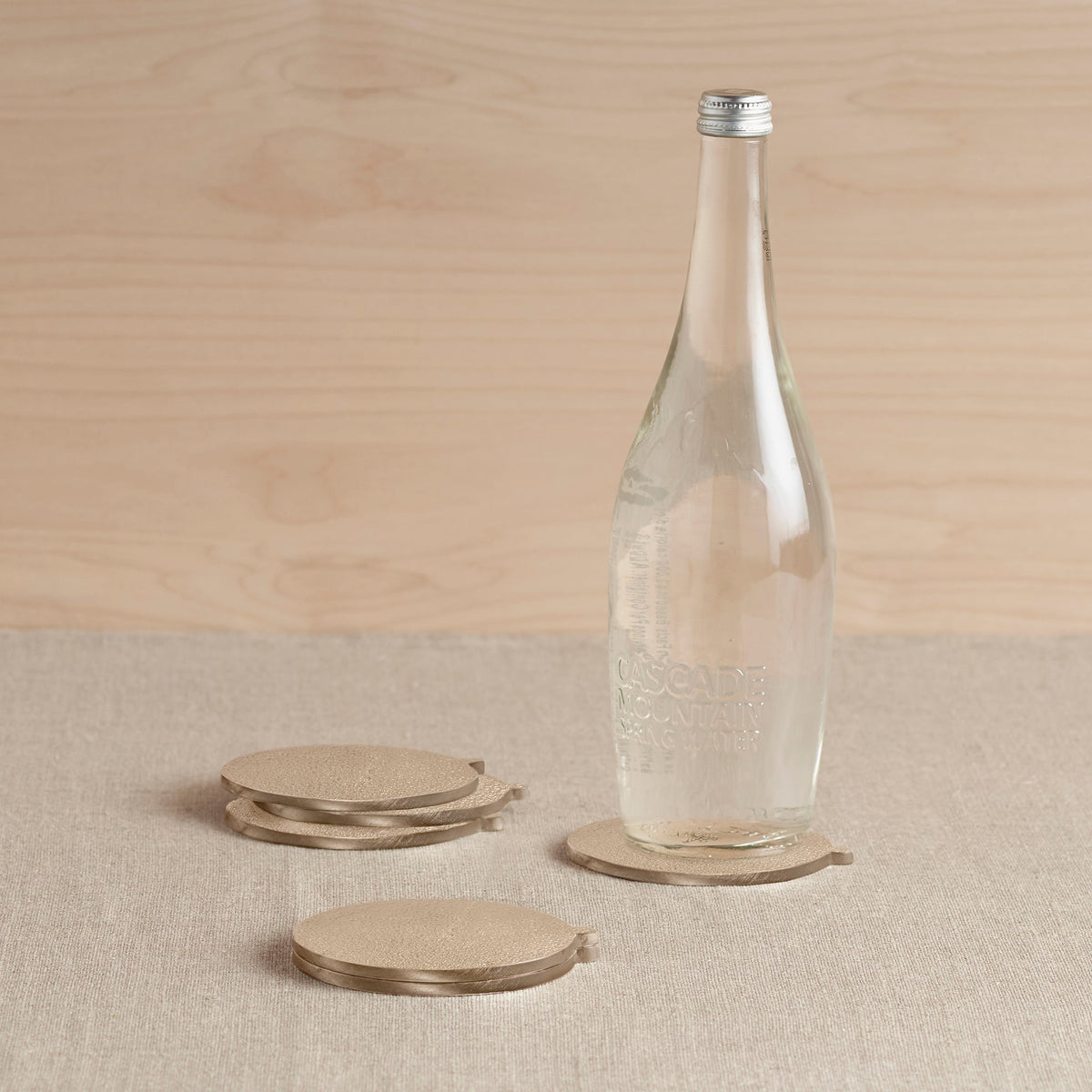Clear glass bottle with metal cap on a beige surface with brass coasters and a wooden background