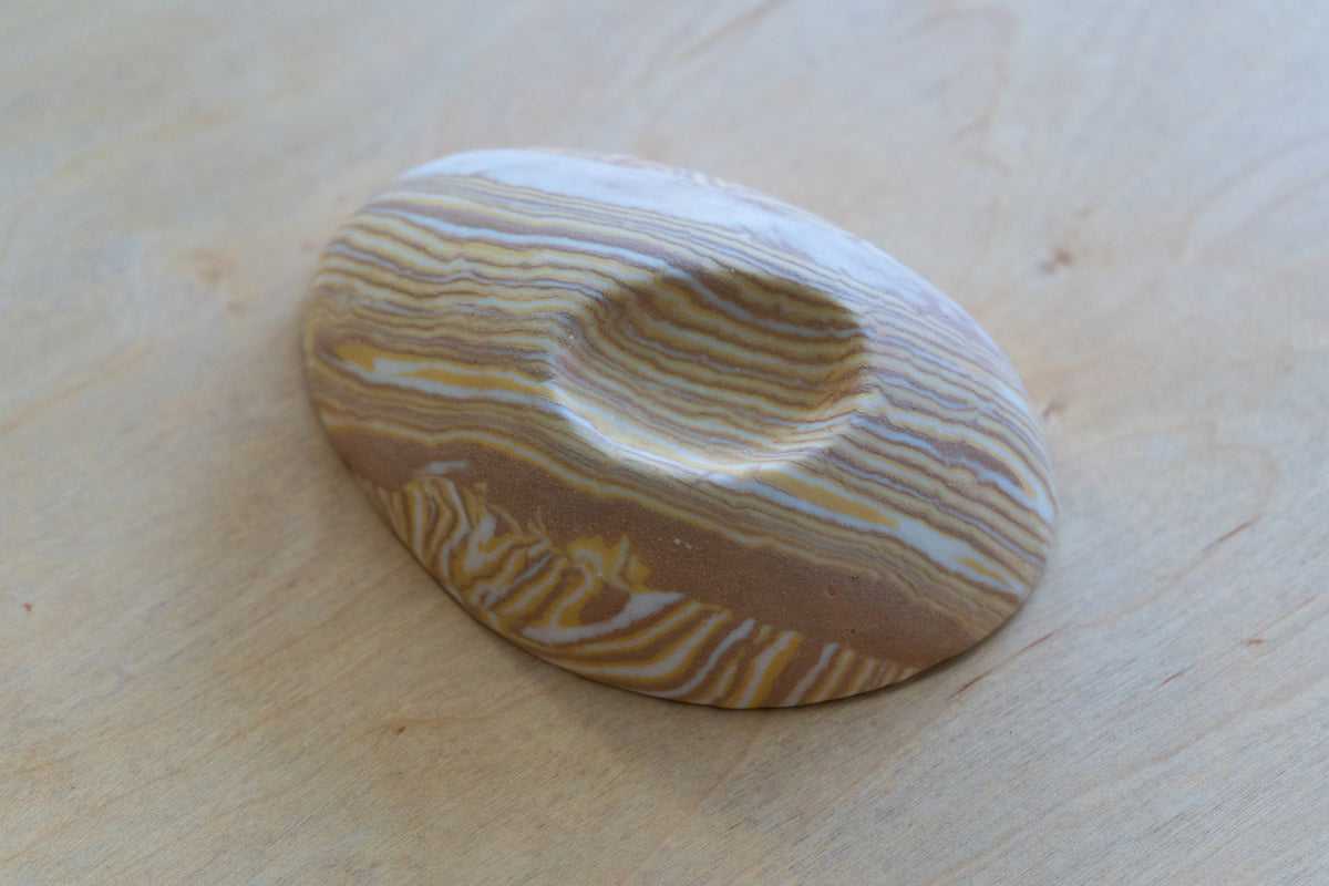 A small oval nerikomi porcelain dish with concentric wavy bands of tan, brown, cream, and pale yellow, photographed on a light wood surface.