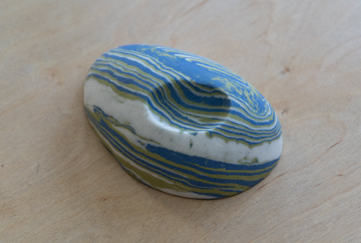 A small oval nerikomi porcelain dish with concentric wavy bands of blue, green, and white, photographed on a light wood surface.