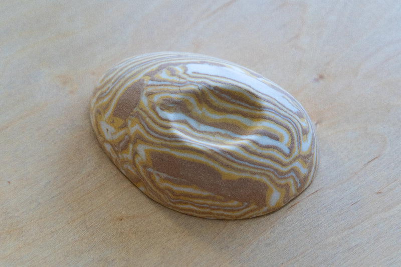 A small oval nerikomi porcelain dish with concentric wavy bands of tan, brown, cream, and pale yellow, photographed on a light wood surface.