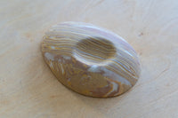 A small oval nerikomi porcelain dish with concentric wavy bands of tan, brown, cream, and pale yellow, photographed on a light wood surface.