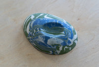 A small oval nerikomi porcelain dish with concentric wavy bands of blue, green, and white, photographed on a light wood surface.