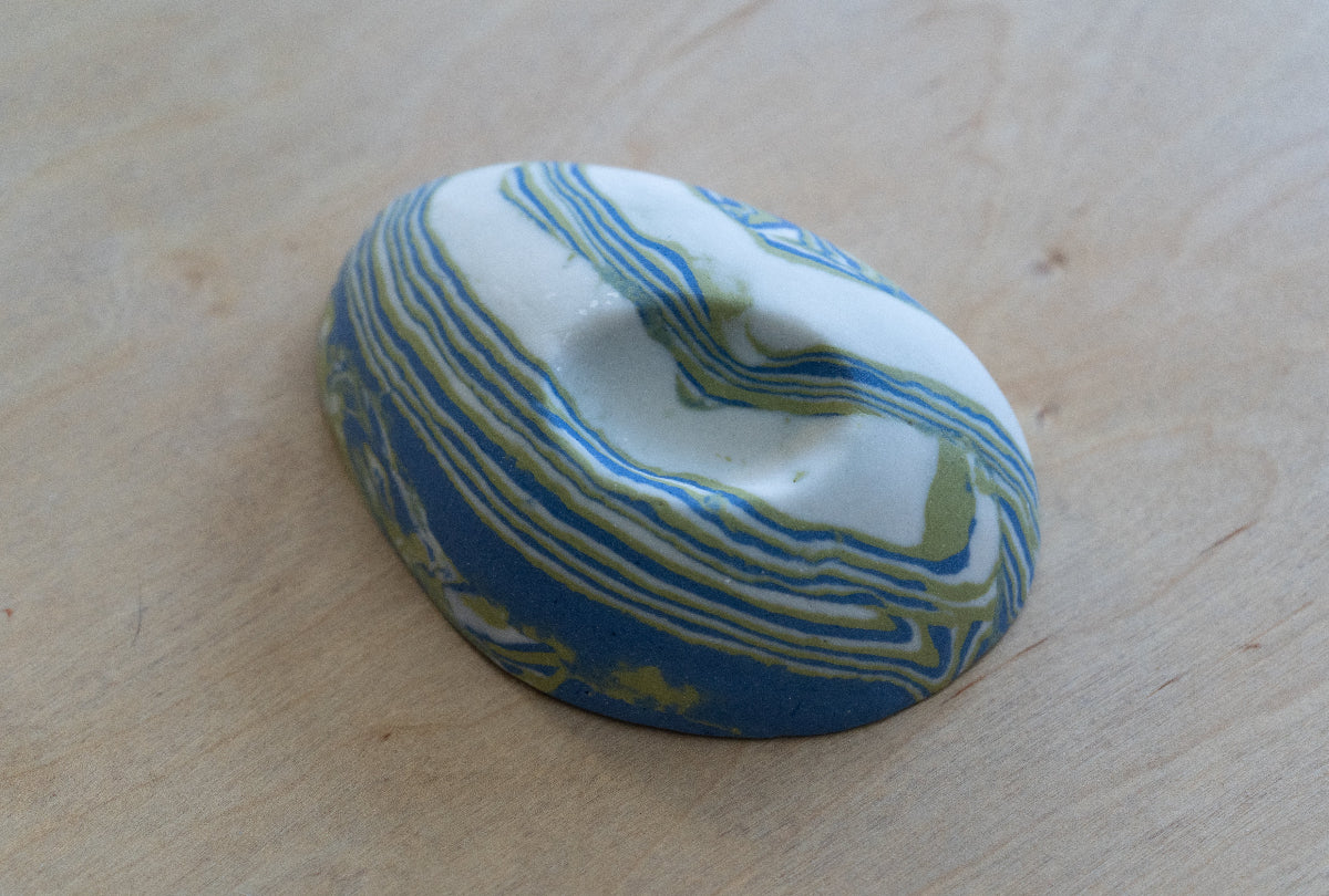 A small oval nerikomi porcelain dish with concentric wavy bands of blue, green, and white, photographed on a light wood surface.