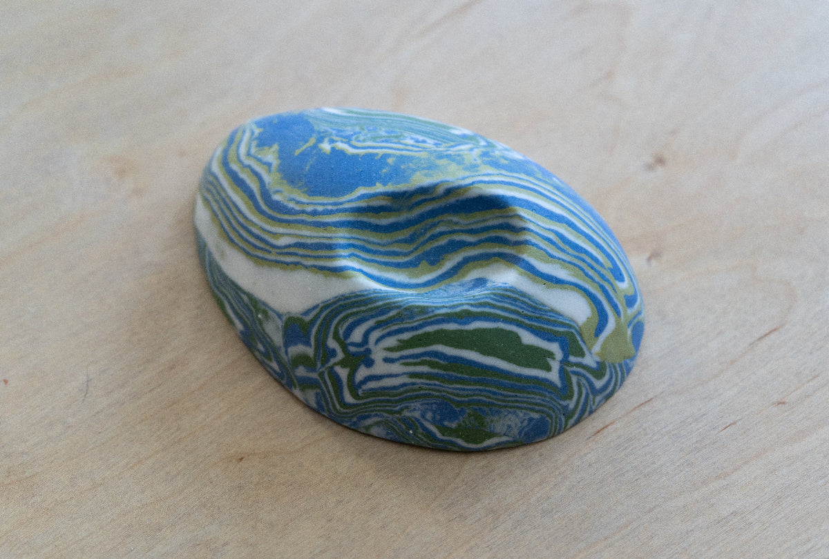 A small oval nerikomi porcelain dish with concentric wavy bands of blue, green, and white, photographed on a light wood surface.