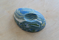 A small oval nerikomi porcelain dish with concentric wavy bands of blue, green, and white, photographed on a light wood surface.
