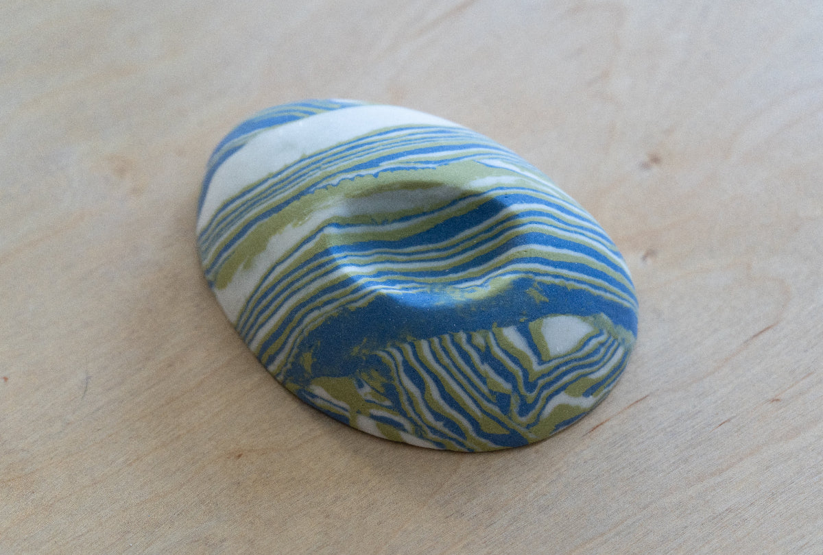 A small oval nerikomi porcelain dish with concentric wavy bands of blue, green, and white, photographed on a light wood surface.