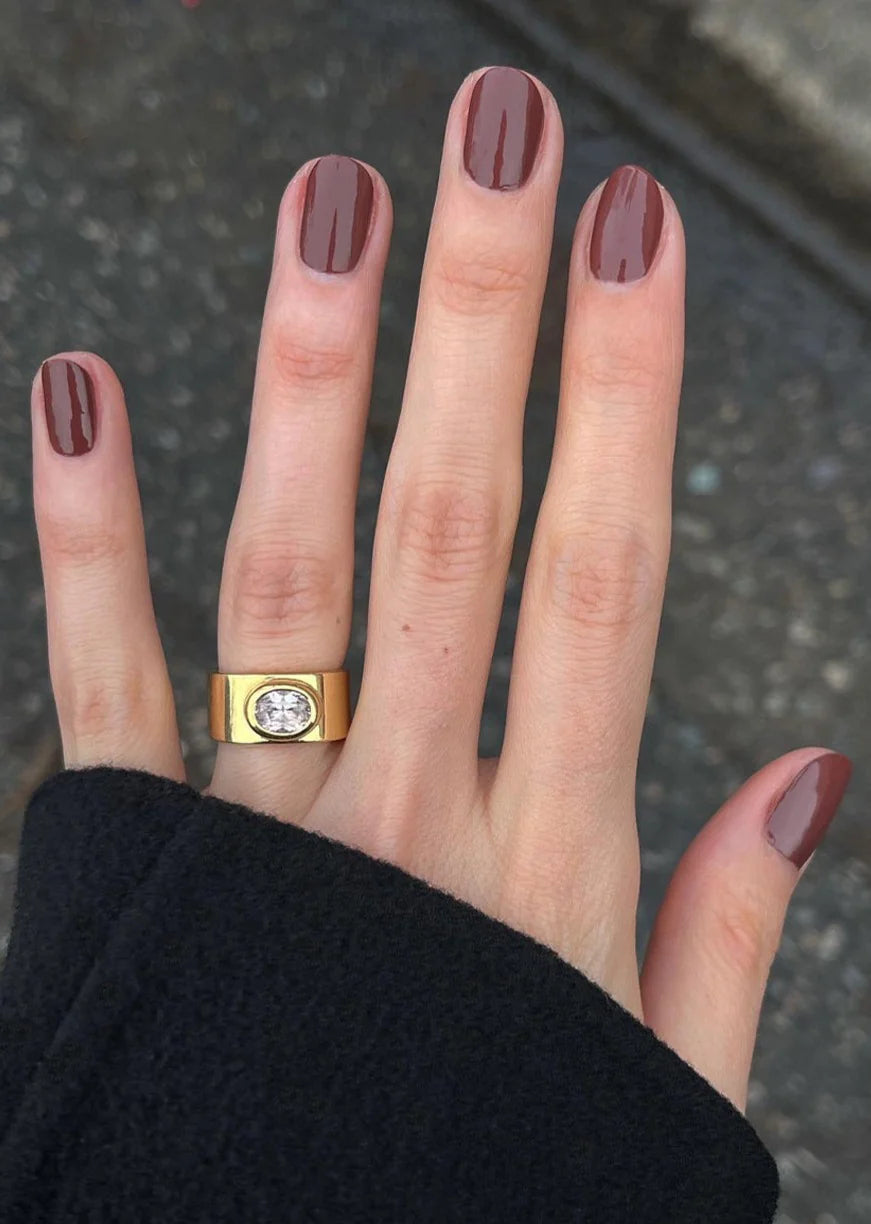 Hand wearing a gold diamond ring and brown nail polish on a dark background