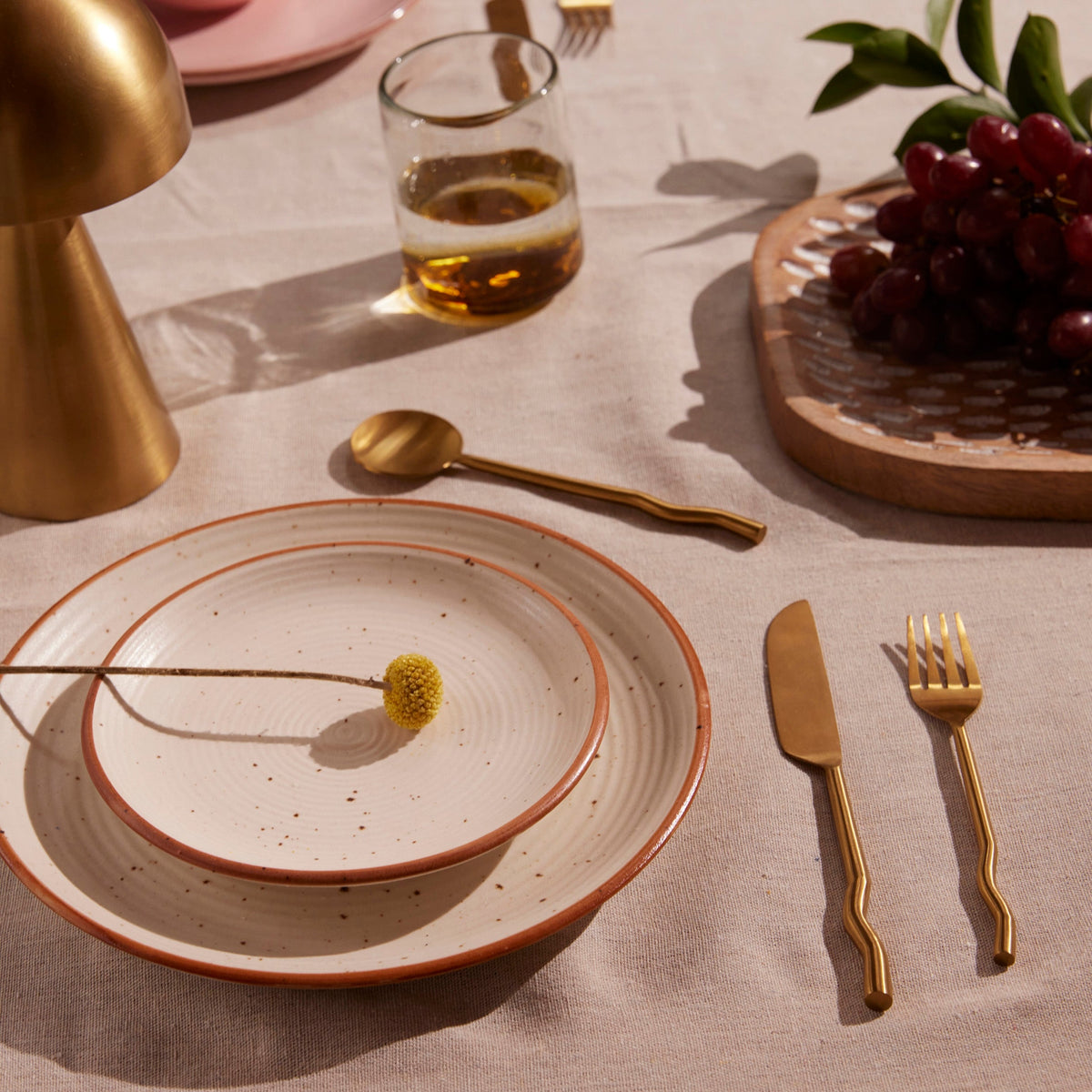 Dinner table setting with brass cutlery, plates, and a glass of wine on a neutral background.
