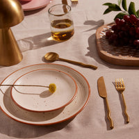 Dinner table setting with brass cutlery, plates, and a glass of wine on a neutral background.