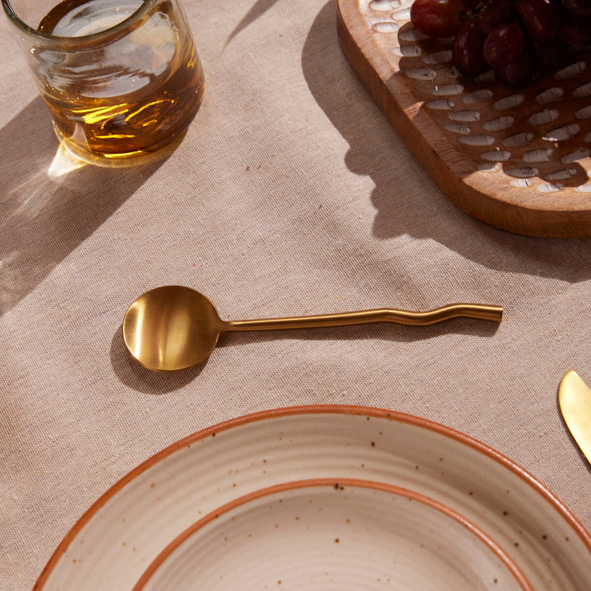 Brass spoon on a textured surface with a glass of amber liquid and a wooden board in the background.