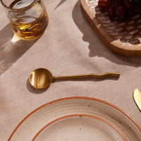 Brass spoon on a textured surface with a glass of amber liquid and a wooden board in the background.