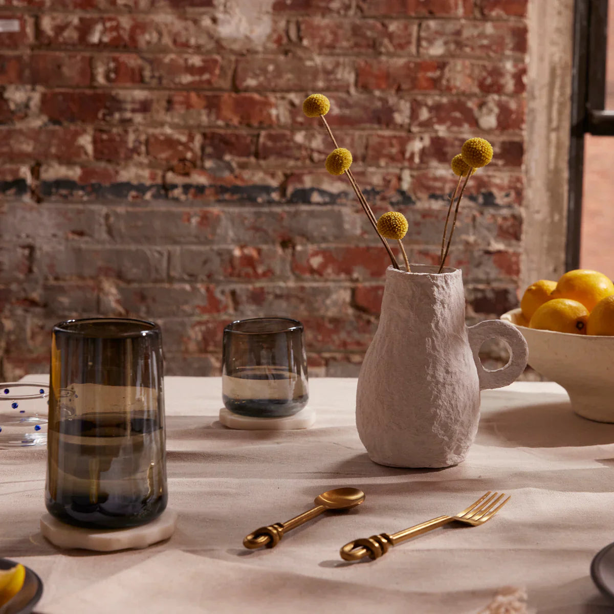 Ceramic vase with dried flowers on a table with glasses and a bowl of lemons against a brick wall.