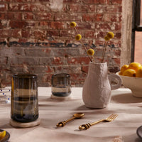 Ceramic vase with dried flowers on a table with glasses and a bowl of lemons against a brick wall.