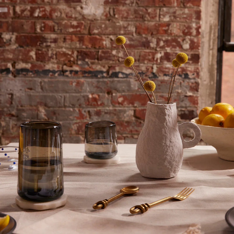 Ceramic vase with dried flowers on a table with glasses and a bowl of lemons against a brick wall.