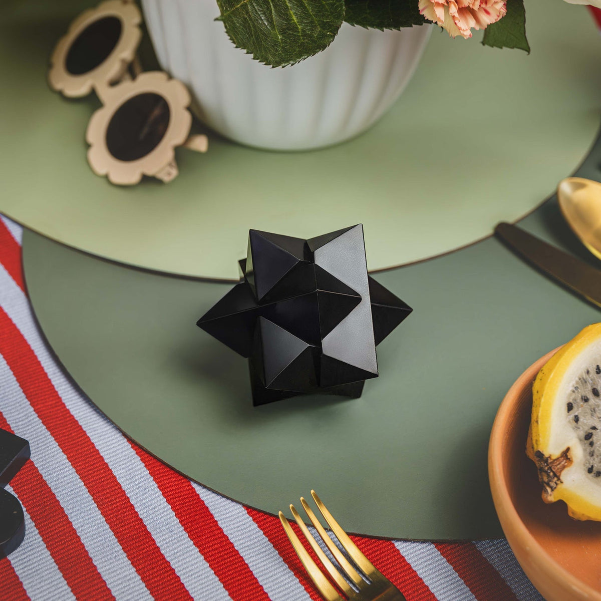 Black geometric object on a green plate with a striped tablecloth and dragon fruit.