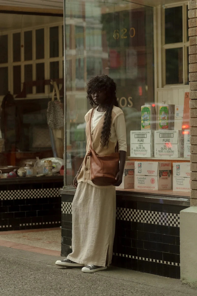 A woman with long dark curly hair stands outside a storefront, wearing a cream linen dress, a tan vest, and carrying a brown leather shoulder bag crossbody.