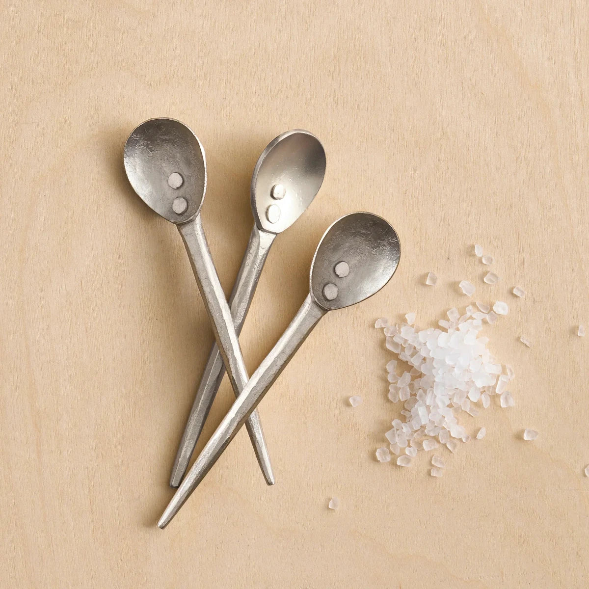 Three pewter mini spoons next to salt on a wooden surface