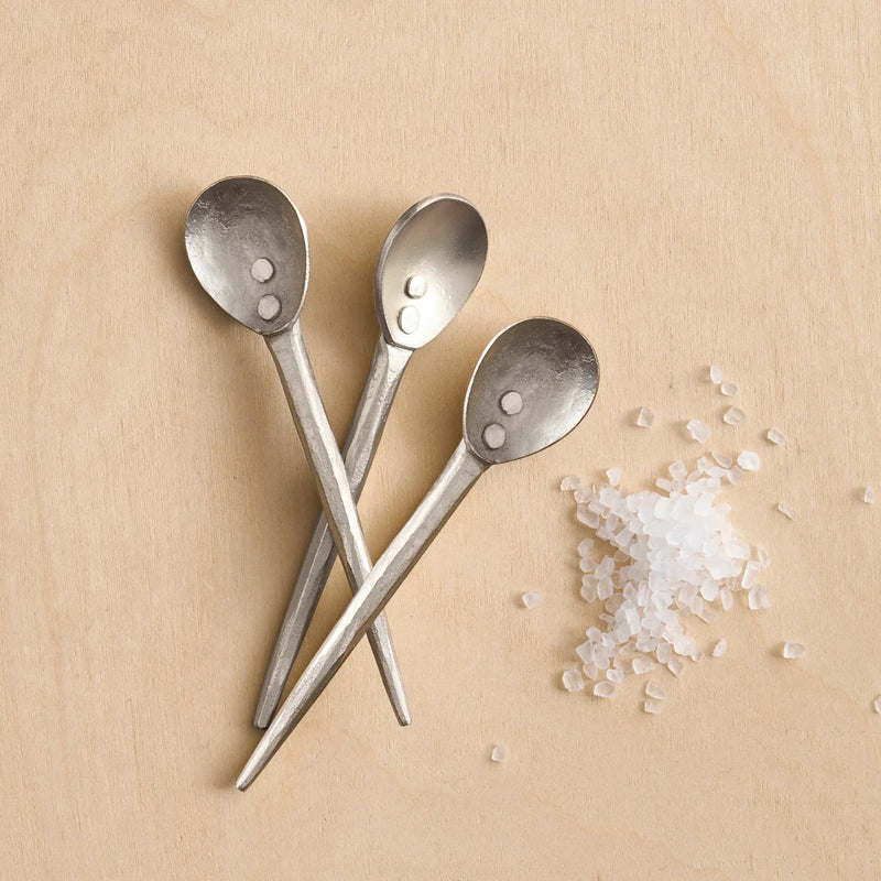 Three pewter mini spoons next to salt on a wooden surface
