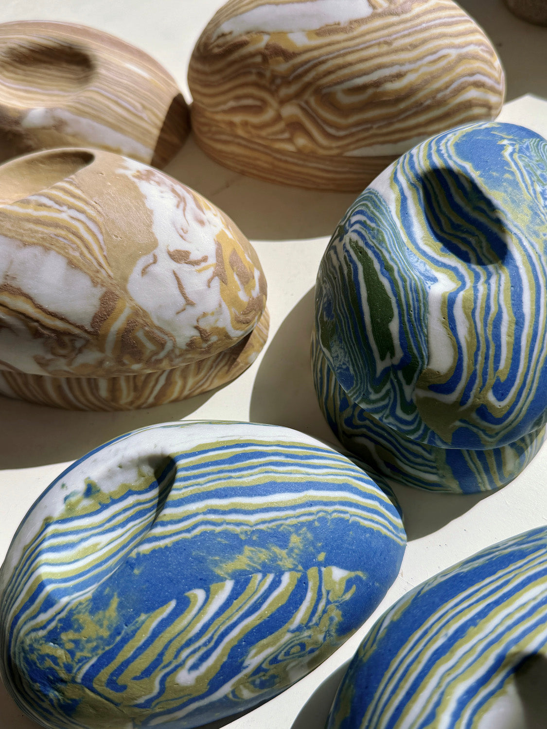 Several small handcrafted pebble shaped vessels with organic marbled nerikomi patterns in blue, white, tan, and olive green, arranged on a light surface.