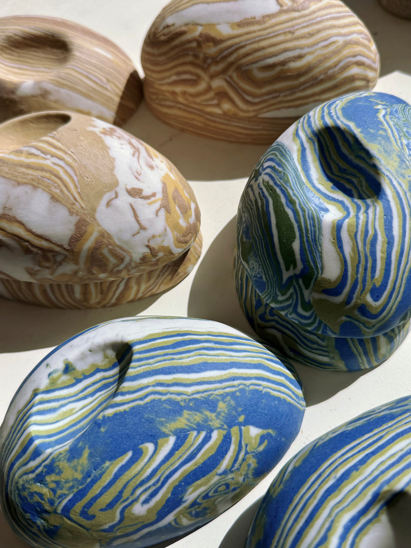 Several small handcrafted pebble shaped vessels with organic marbled nerikomi patterns in blue, white, tan, and olive green, arranged on a light surface.