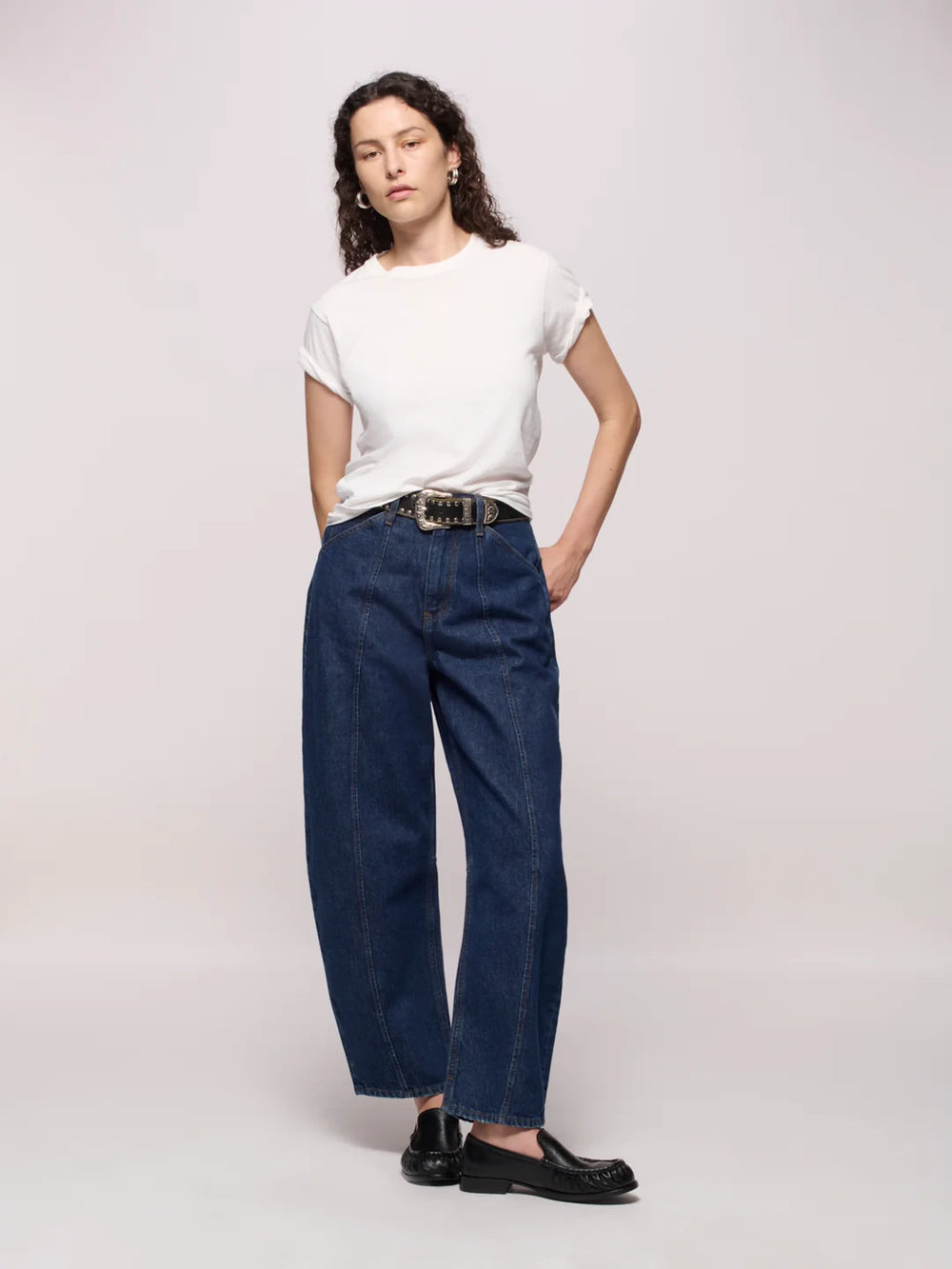 Woman wearing dark blue jeans belted, a white tee, and black loafers with silver hoop earrings in front of a white background