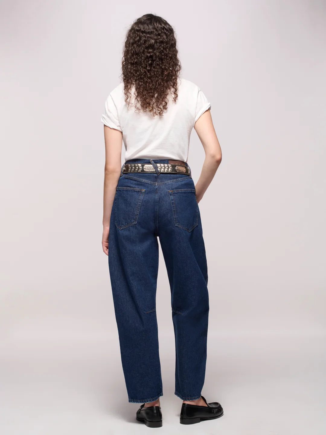 Backside of a person wearing dark blue jeans belted, a white tee, and black loafers in front of a white background
