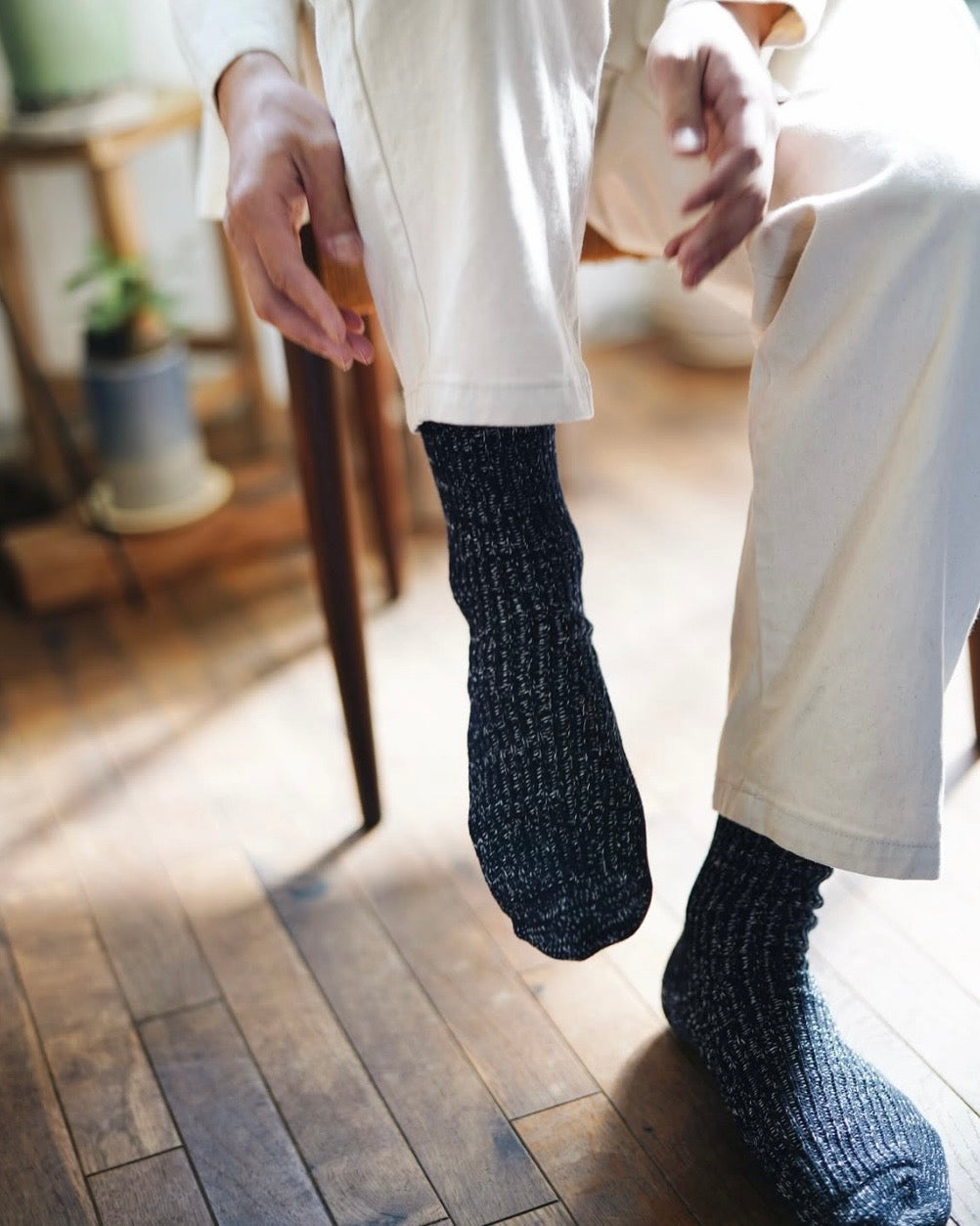 person sitting in a chair with one foot raised and one foot flat on the floor, reaching toward their foot wearing speckled navy blue socks and white pants on a wooden floor with blurry furniture in the background