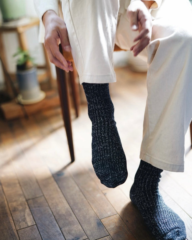 person sitting in a chair with one foot raised and one foot flat on the floor, reaching toward their foot wearing speckled navy blue socks and white pants on a wooden floor with blurry furniture in the background