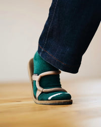 Person's foot taking a step wearing green socks with a white toe stripe, tan sandals, and dark denim pants on a light wooden floor
