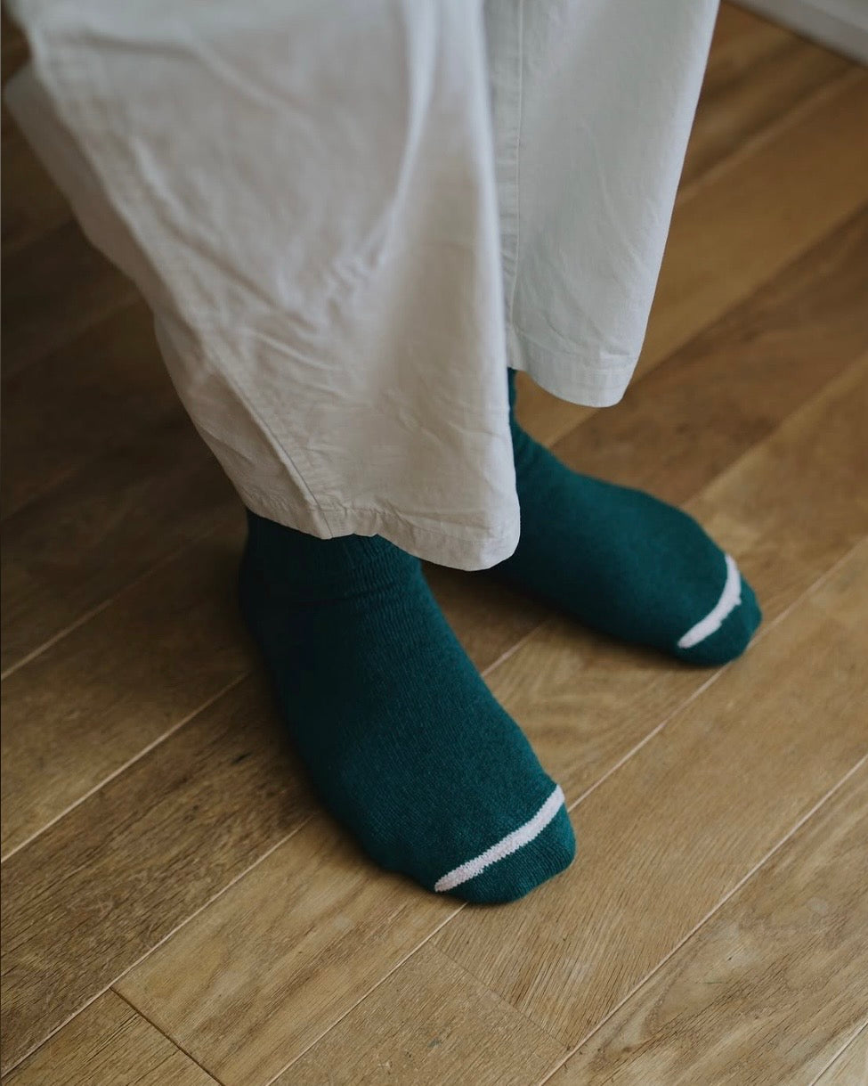 View looking down on person's feet wearing dark green socks with a white toe stripe and beige pants on a light wooden floor