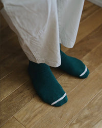 View looking down on person's feet wearing dark green socks with a white toe stripe and beige pants on a light wooden floor