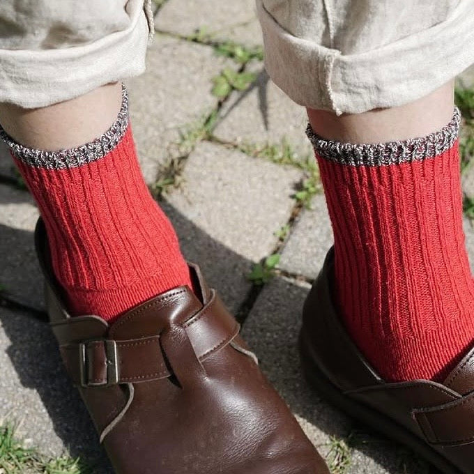 Closeup of person's ankles wearing red ribbed socks with brown leather clogs and cuffed beige pants on a brick ground surface