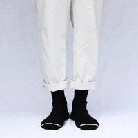 Person wearing black ankle socks with a white toe stripe and cuffed white pants on a light grey background