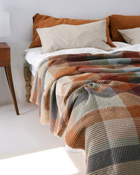 A textured waffle-knit blanket with a warm plaid pattern in rust, beige, brown, and muted blue-gray, styled on a bed with matching pillows in a bright, minimalist bedroom.