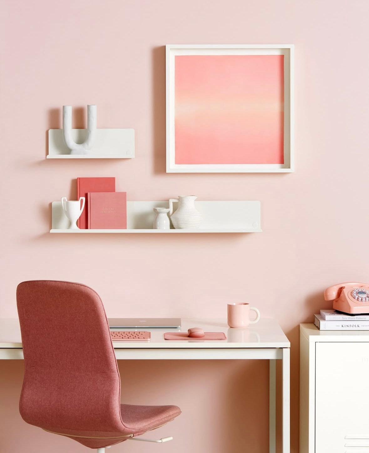 Modern home office with pink chair, desk, decorative shelves, and wall art on a light pink wall.