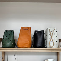 Four leather bags in green, brown, black, and white on a wooden shelf against a plain wall.