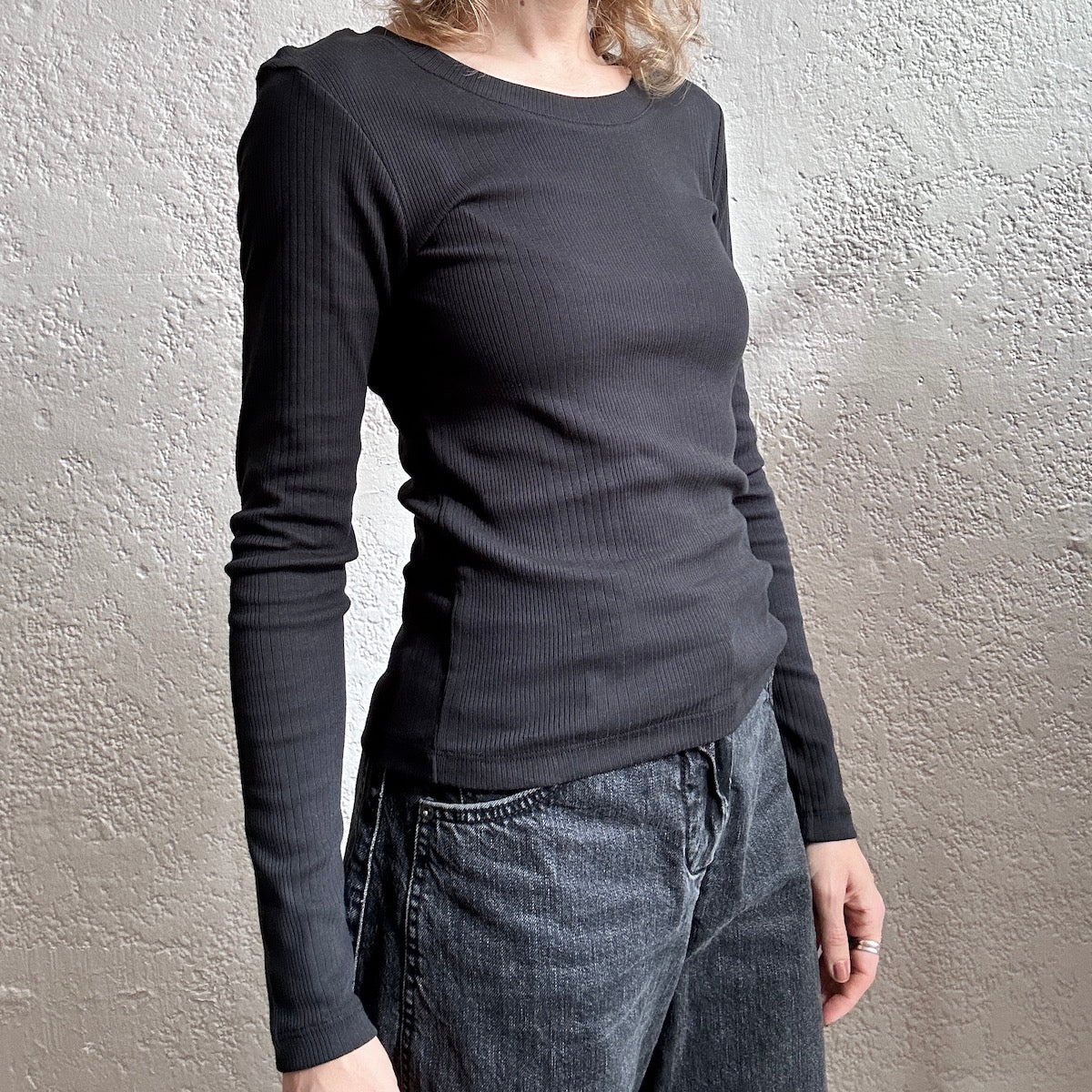 Person wearing a black long-sleeve top and jeans against a neutral wall.