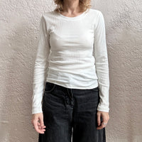 Person wearing a white long-sleeve shirt and dark jeans against a plain wall.
