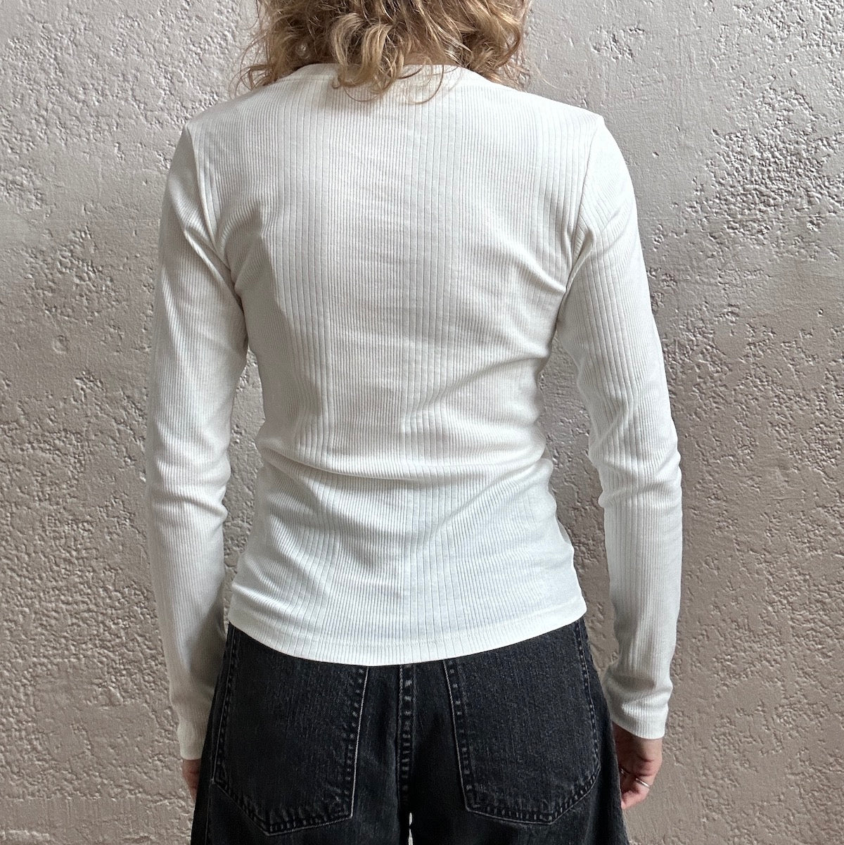 Person wearing a white ribbed long-sleeve shirt against a textured gray wall.