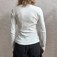 Person wearing a white ribbed long-sleeve shirt against a textured gray wall.