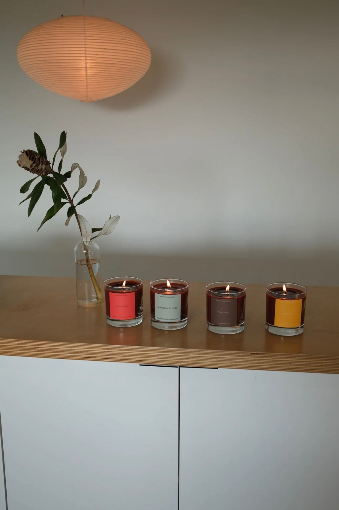 Four candles with different colored labels on a wooden surface, with a vase of flowers and a lamp in the background.