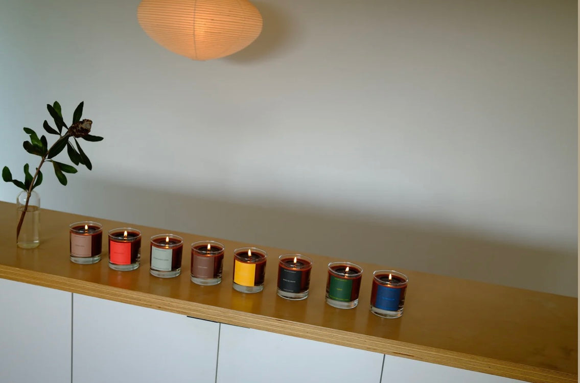 Row of colorful candles on a wooden shelf with a plant and light fixture in the background.