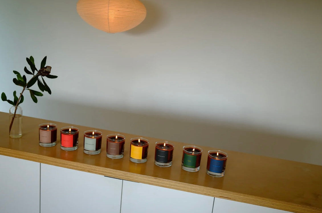 Row of colorful candles on a wooden shelf with a plant and light fixture in the background.