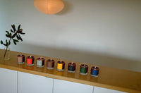 Row of colorful candles on a wooden shelf with a plant and light fixture in the background.