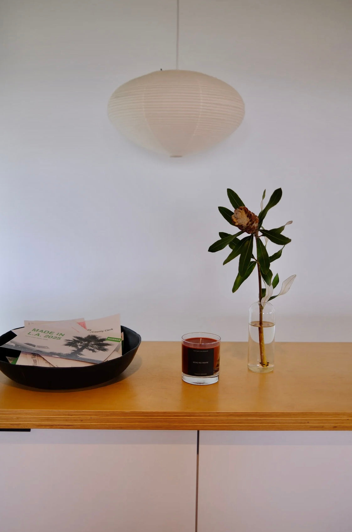 Small table with a glass jar candle, a bowl with books, and a plant against a white wall.