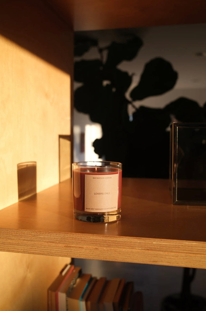 Candle with a label on a wooden shelf with books and a plant in the background
