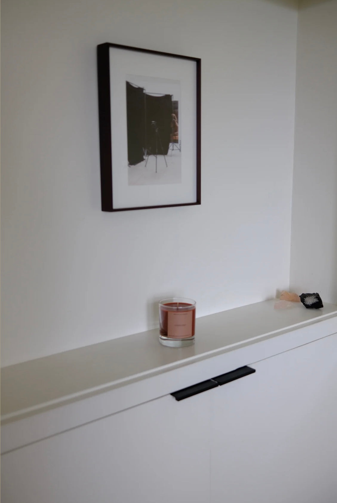 Framed picture on a white wall above a shelf with a glass and small object.