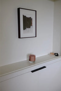 Framed picture on a white wall above a shelf with a glass and small object.